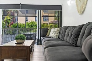 A living room with a dark grey sofa, a wooden coffee table with a plant, and a sliding glass door leading to a balcony with greenery and a building in the background.