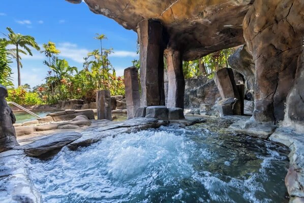 A natural hot spring with flowing water, surrounded by large rock formations and tropical vegetation.