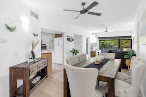 A spacious dining area with a wooden table and white chairs, a kitchen in the background, and a ceiling fan.