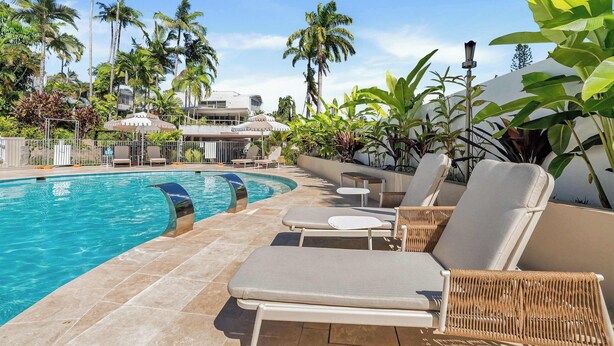 Un área junto a la piscina con sillones de descanso, una pileta, palmeras y un edificio al fondo.