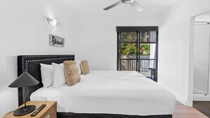 Un dormitorio con cama, una mesita de noche con lámpara, un ventilador de techo, una ventana con vistas a zonas verdes y un cartel en la pared.