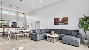 Ein modernes Wohnzimmer mit einem grauen Ecksofa, einem runden Couchtisch und einem Essbereich mit weißem Tisch und Stühlen.