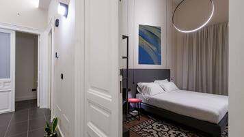 Ein modernes Hotelzimmer mit einem Bett, einem Schreibtisch, einem Sessel, einer Topfpflanze und einem großen, abstrakten Gemälde an der Wand.
