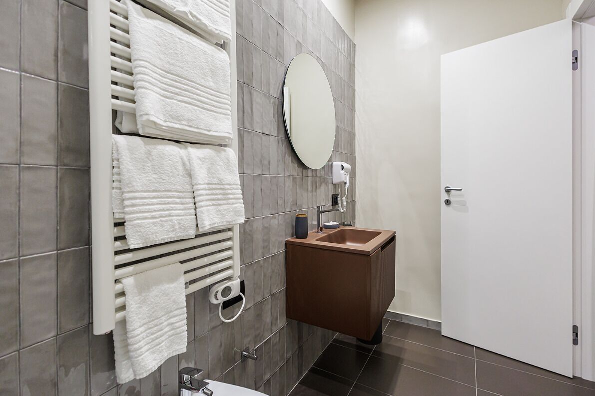 Une salle de bain avec des murs recouverts de carreaux gris, un porte-serviettes blanc avec des serviettes pliées, un miroir rond, un lavabo marron et une porte blanche.