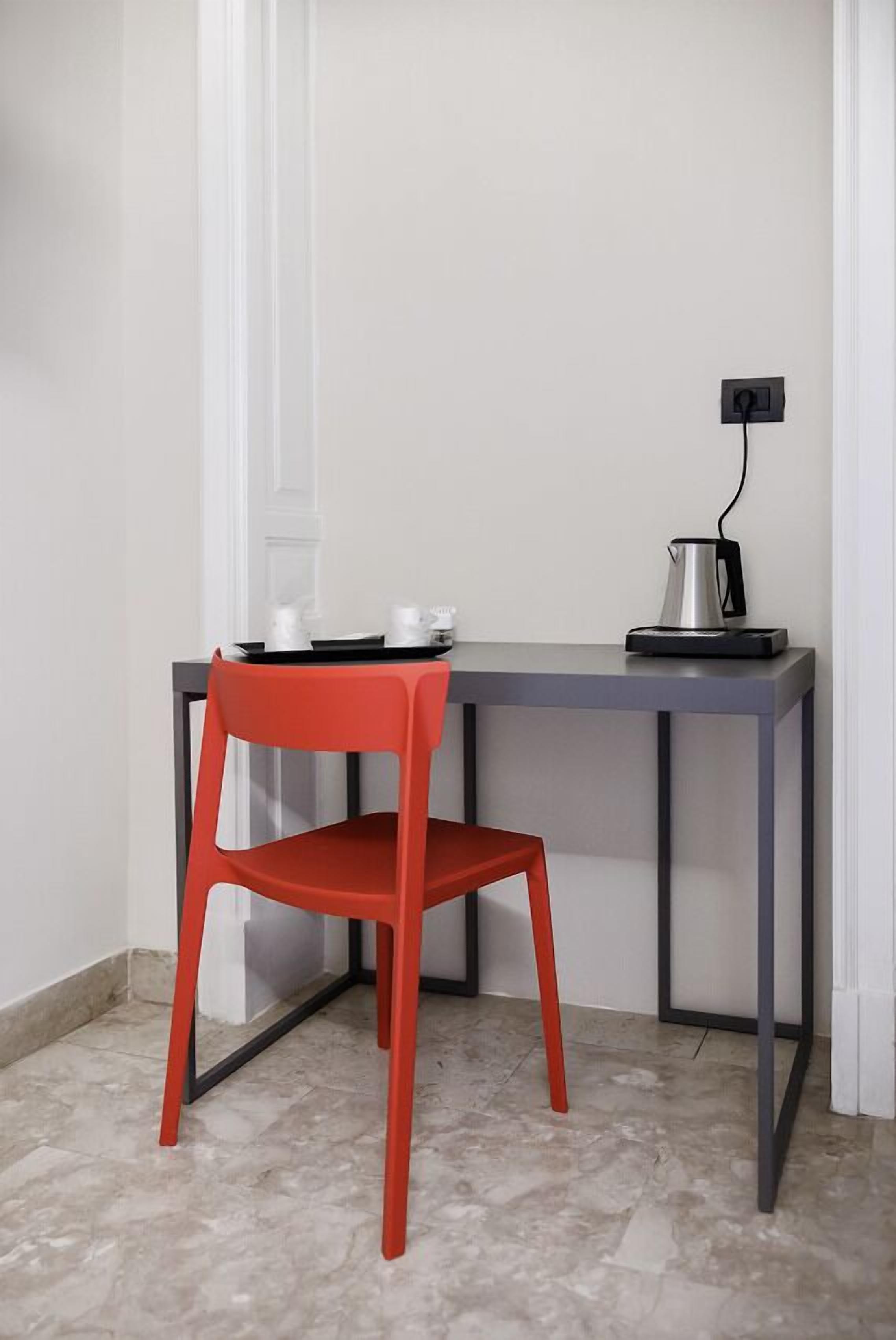 A small, modern room with a red chair and a grey table. There is a kettle on the table and a power outlet on the wall.