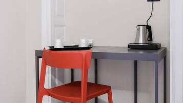 A small, modern room with a red chair and a grey table. There is a kettle on the table and a power outlet on the wall.