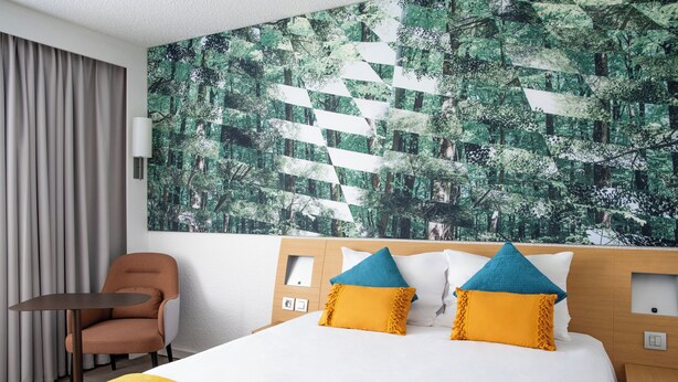 A hotel room with a bed, a chair, a table, and a large wall mural. - Sweet Home By Novotel Bordeaux Mérignac (Mérignac)