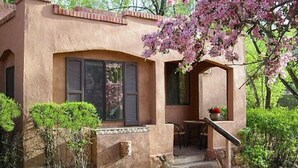 Una casa de estuco con un porche cubierto, un pequeño espacio para sentarse al aire libre y un jardín con un árbol en flor.