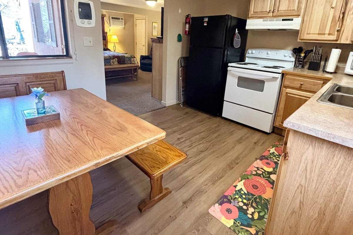 A compact kitchen with wooden cabinets, a black refrigerator, a white stove, and a dining area with a wooden table and bench.