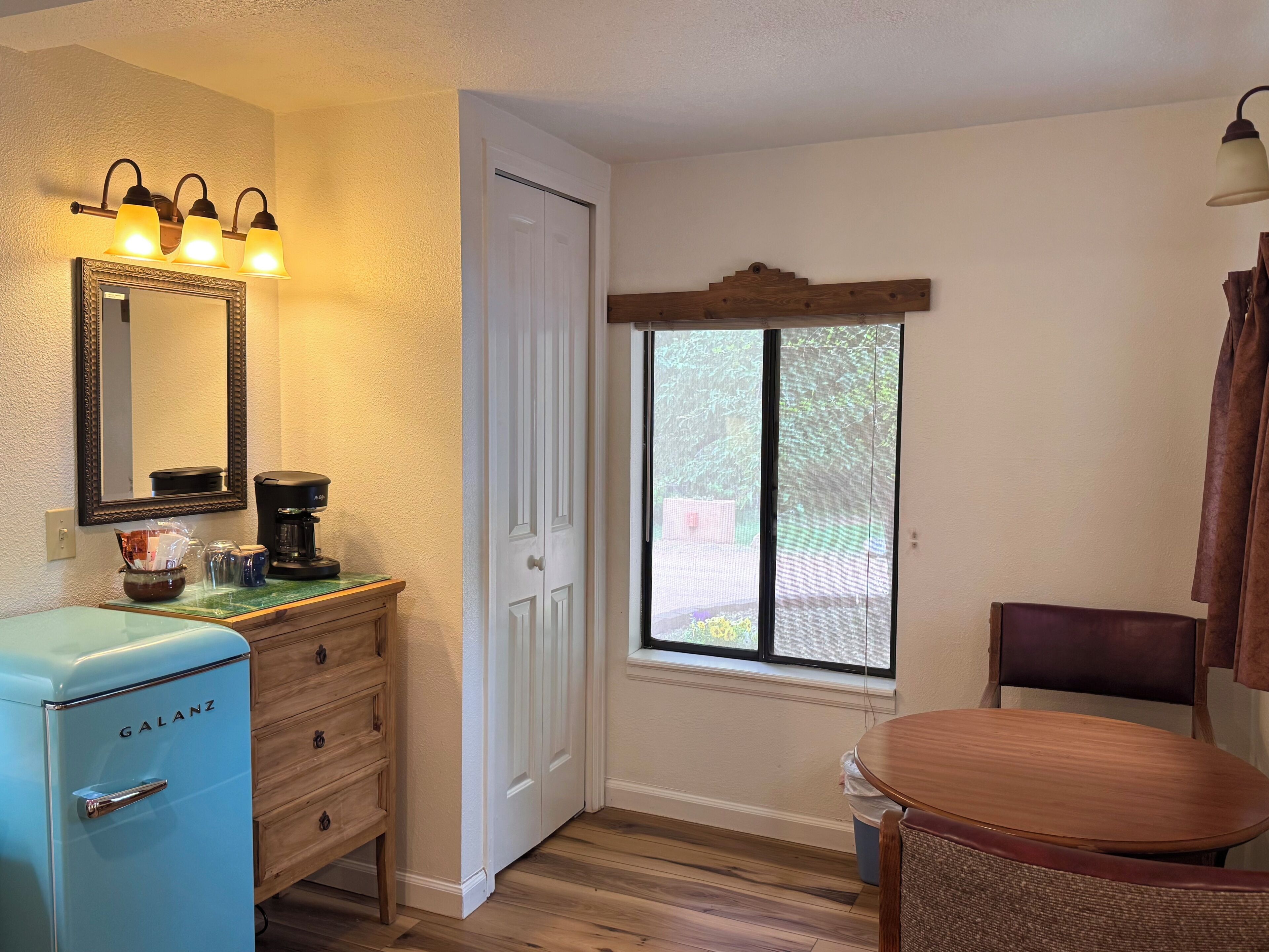 A room with a vintage blue refrigerator, a coffee maker, a mirror, a door, a window, and a chair.