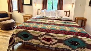 Un dormitorio con cama, dos mesitas de noche, una silla y un ventanal con cortinas.