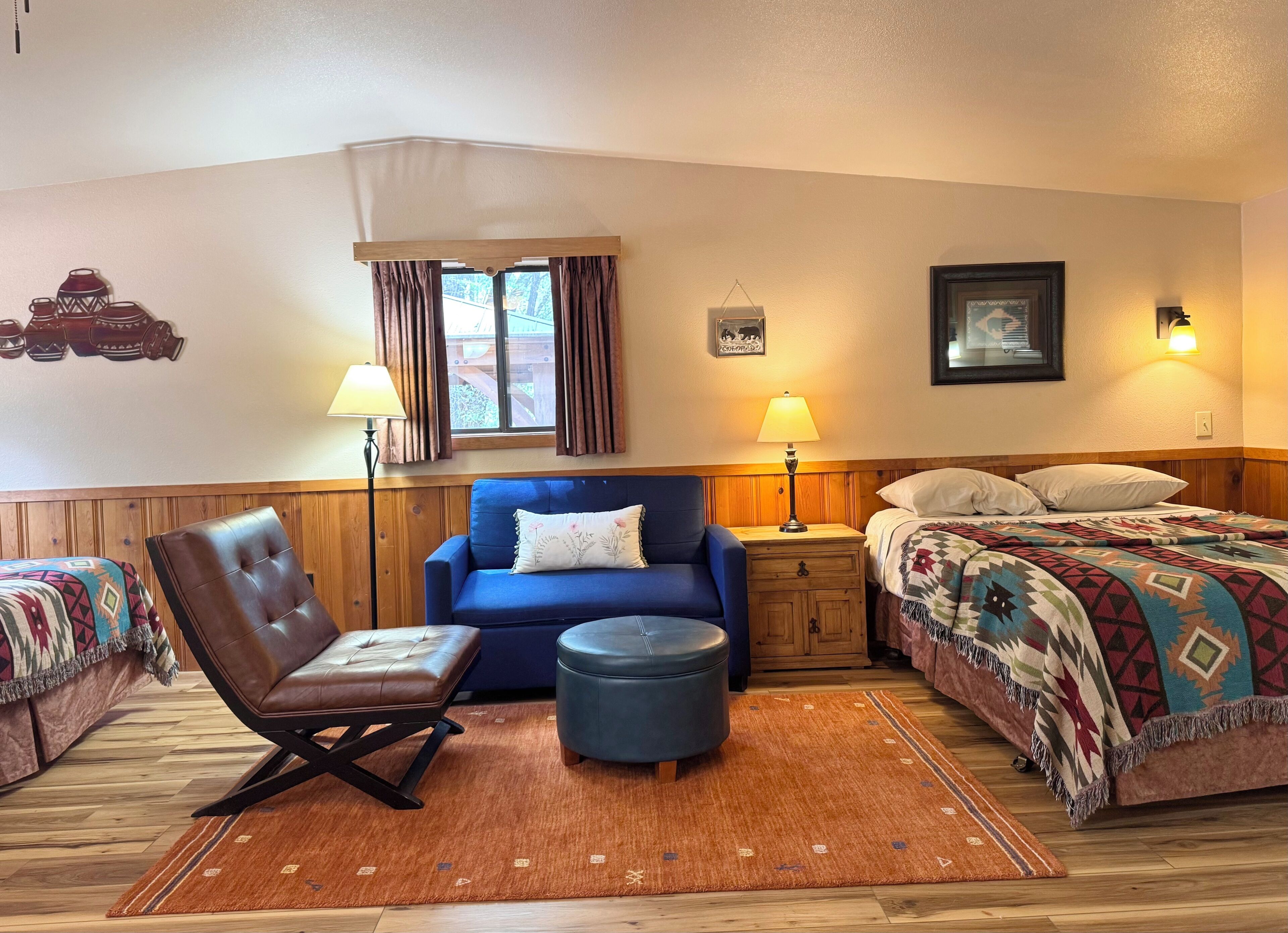 A room with a bed, a blue sofa, a brown leather chair, a wooden nightstand, a lamp, and a window with curtains.