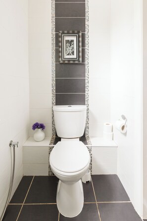 Un baño con un inodoro blanco, un estante pequeño con un jarrón de flores moradas y un cuadro en la pared.