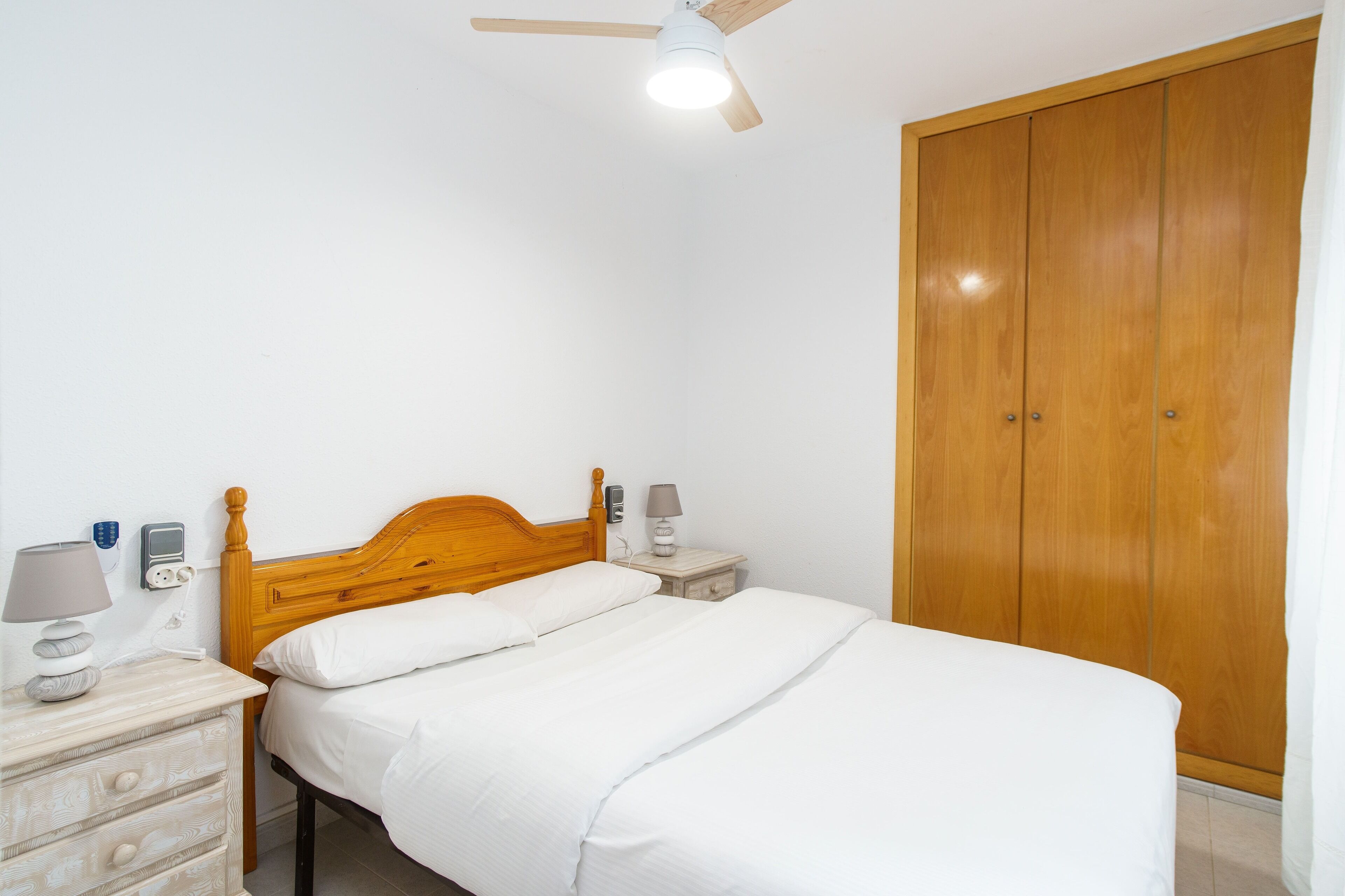 A bedroom with a wooden bed, bedside tables, a nightstand, a ceiling fan, and a wardrobe.