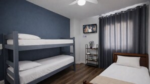 Un letto a castello con biancheria bianca, un letto singolo con biancheria bianca, una testiera in legno, una ventola a soffitto, un televisore a parete e uno scaffale con libri e una pianta.