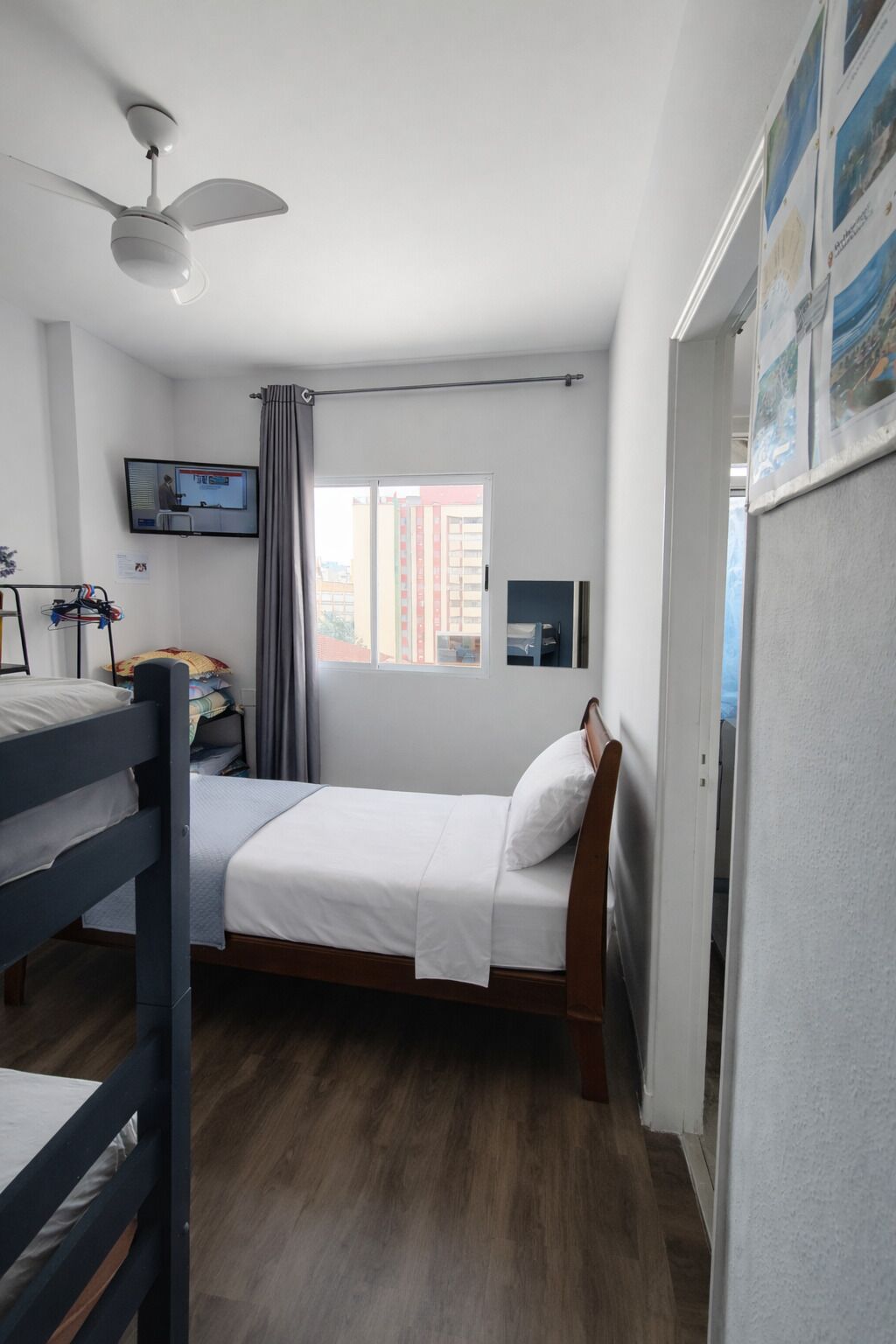A single bedroom with a bed, a ceiling fan, a TV, a window with curtains, and a view of buildings outside.