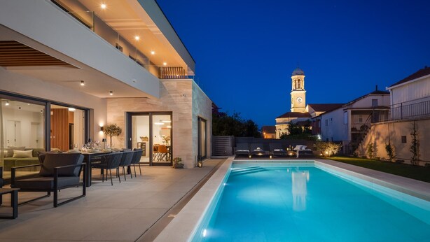 A modern house with a swimming pool, a dining area, and a view of a church with a bell tower. - Villa Prima Lux (Kastela)