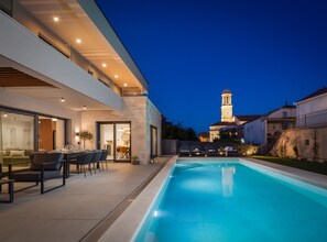 A modern house with a swimming pool, a dining area, and a view of a church with a bell tower. - Villa Prima Lux (Kastela)
