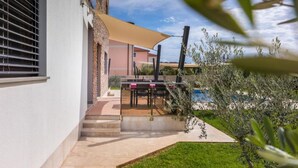 A patio area with a table and chairs, a pool, and a building with a beige awning. - Casa Gigi (Medulin)
