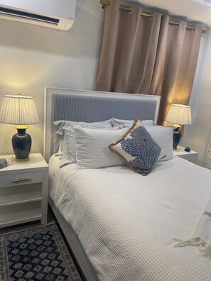 Een opgemaakt bed met witte lakens en decoratieve kussens, met aan beide kanten nachtkastjes met lampen en een decoratieve vaas.