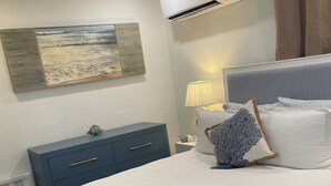 A neatly made bed with a blue and white blanket, a bedside lamp, and a wall-mounted air conditioner. - Siesta Key Beach Cottage-Prime Location! Walkable. Luxury Amenities & Comfort! (Sarasota)