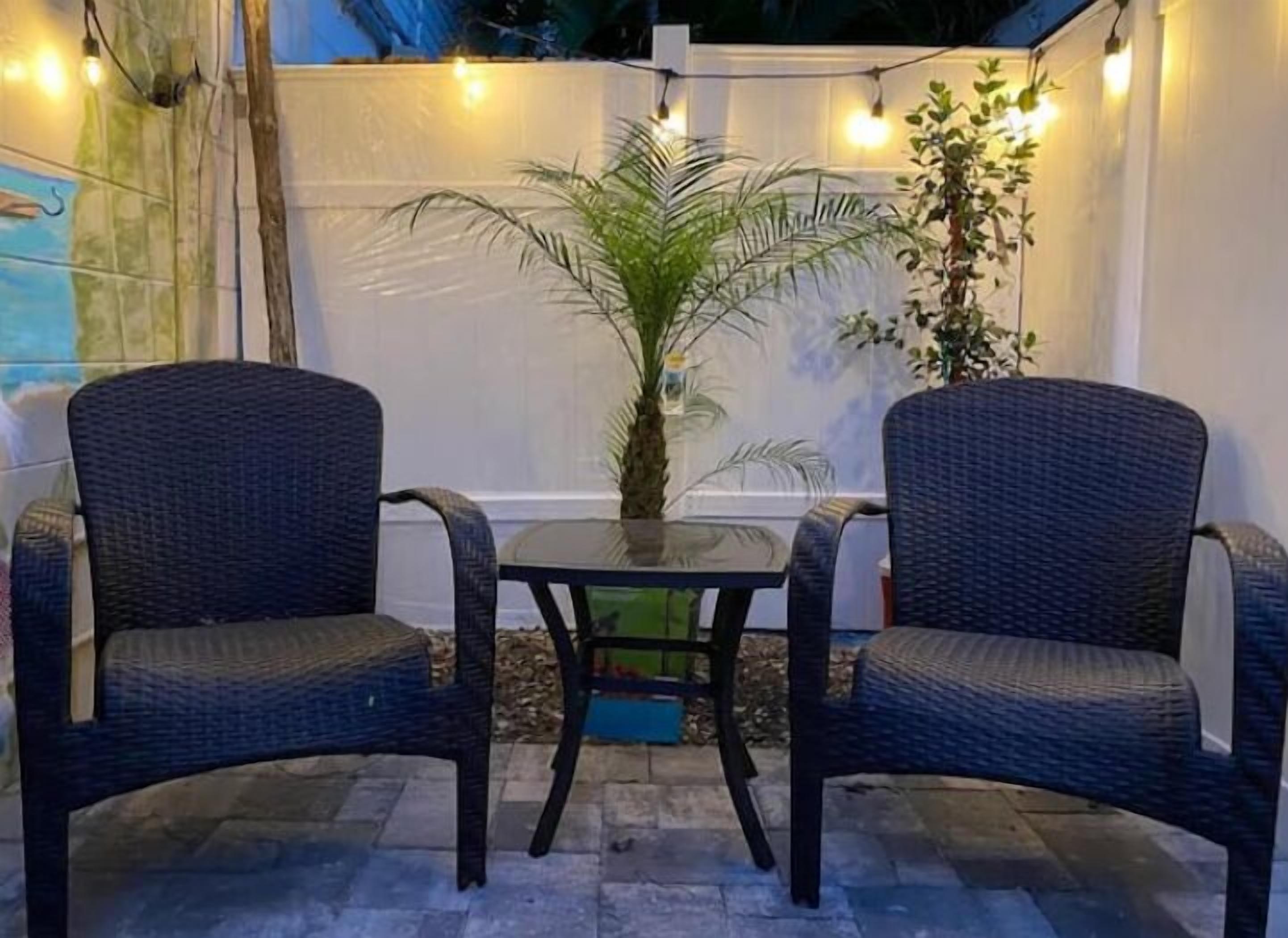 Deux fauteuils en osier sont placés face à une petite table dont le plateau est en verre, le tout étant entouré de guirlandes lumineuses et de plantes.