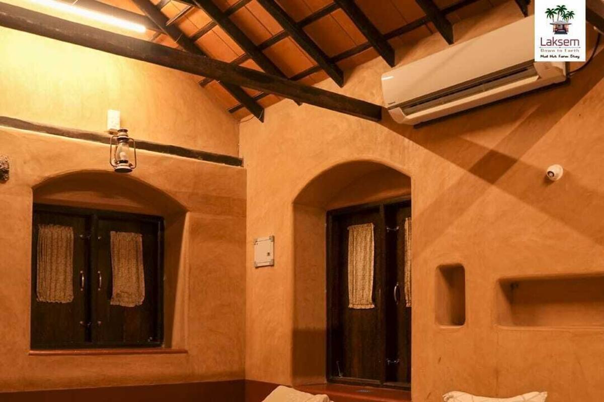 A hotel room with two single beds, a bench, a window with curtains, an air conditioning unit, and a wall-mounted light fixture.