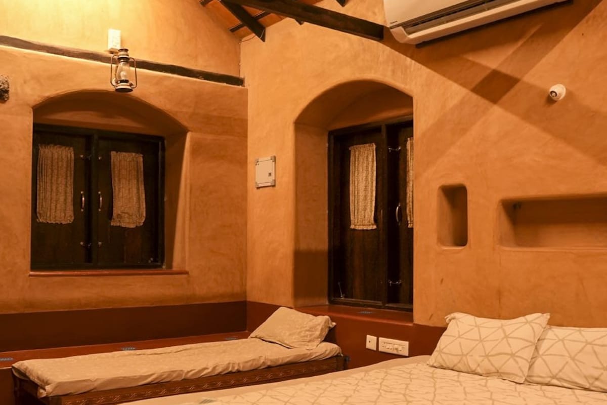 A hotel room with two single beds, a bench, a window with curtains, an air conditioning unit, and a wall-mounted light fixture.