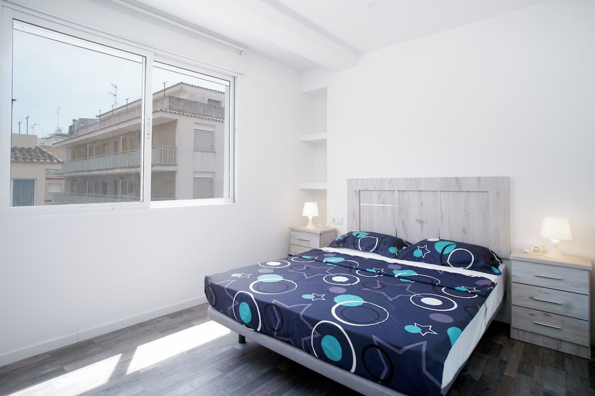 A bedroom with a bed, two bedside tables, a window, and a view of buildings outside.