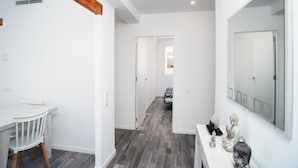 Un corridoio con parete bianca, trave in legno a soffitto, tavolo con sedie, pavimento in legno e uno specchio sulla parete destra.
