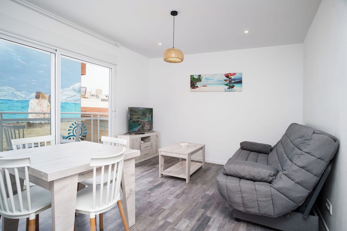 A modern living room with a large window overlooking a beach, a grey sofa, a small table, and a TV.