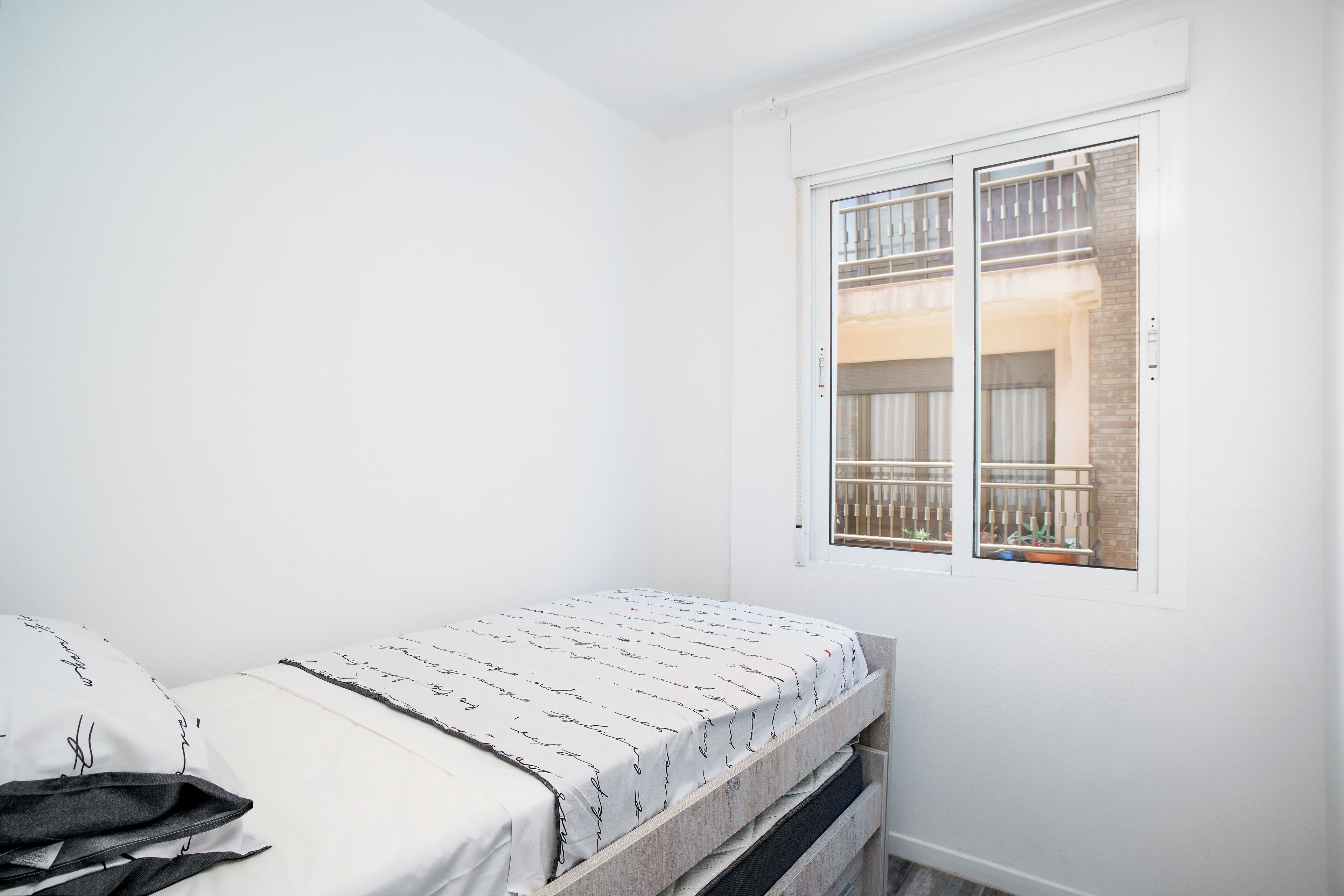 A small, white room with a single bed, a window with a view of a balcony, and a door.