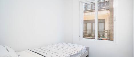 A small, white room with a single bed, a window with a view of a balcony, and a door.