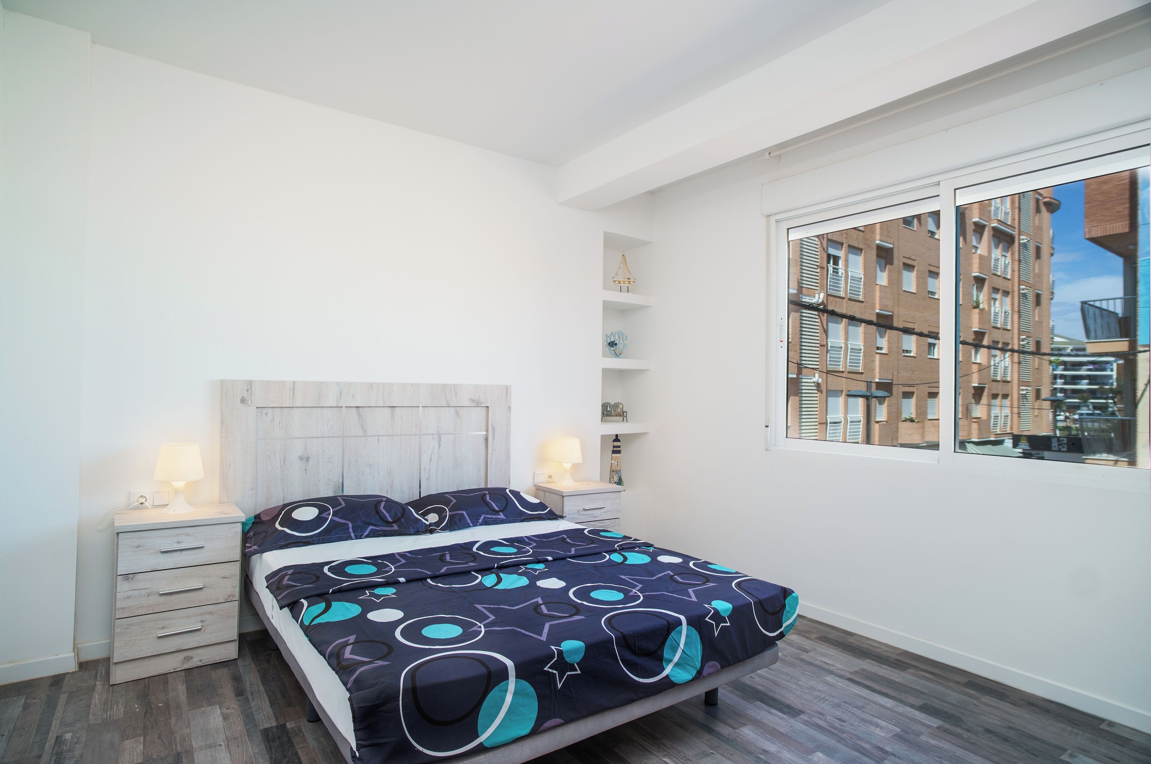 A bedroom with a bed, nightstands, a dresser, a window with a city view, and a wooden headboard.