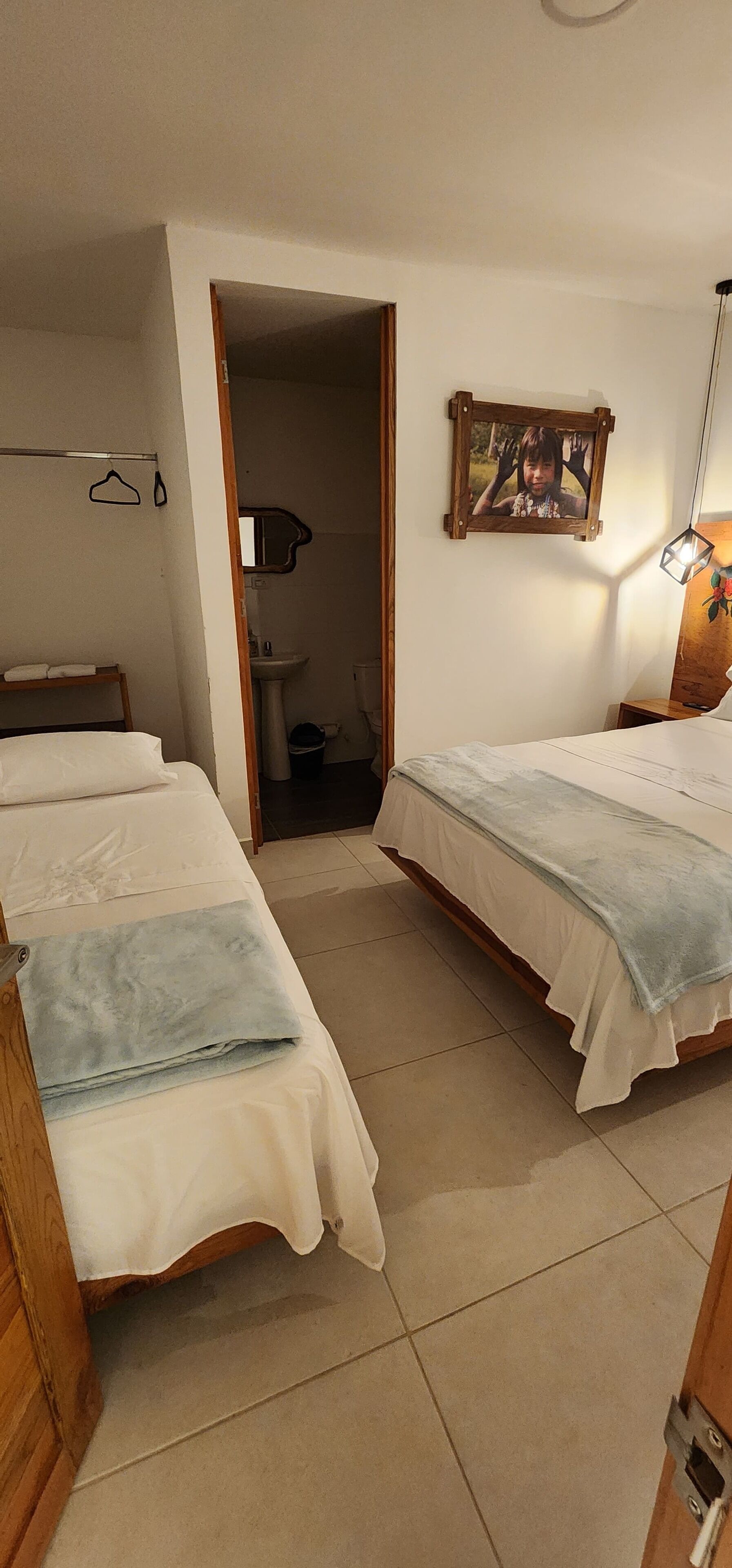 A hotel room with two single beds, a wooden wardrobe, a bedside table with a lamp, a picture on the wall, and a bathroom visible through an open door.