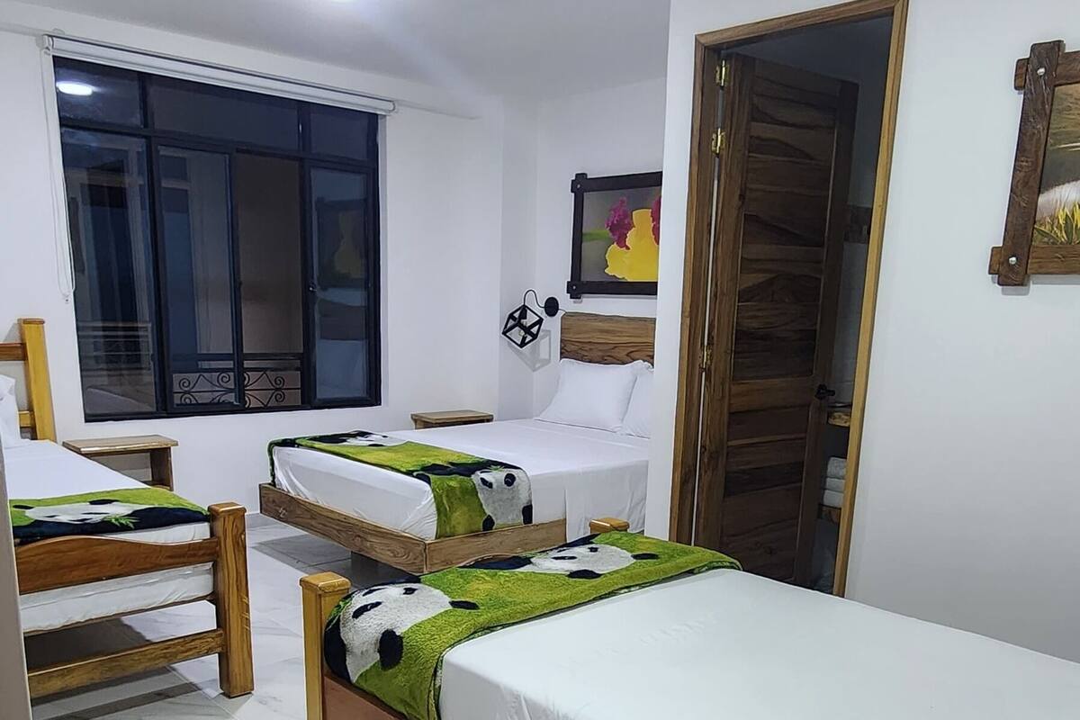 A hotel room with three single beds, a wooden wardrobe, a large window with blinds, and a ceiling light.