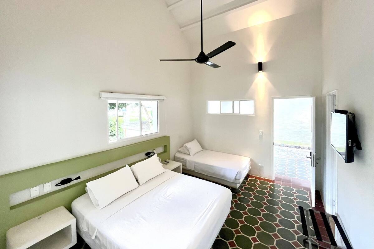 Habitación de hotel con una cama grande, una cama más pequeña, un ventilador de techo, una ventana y una puerta que da a un área exterior.