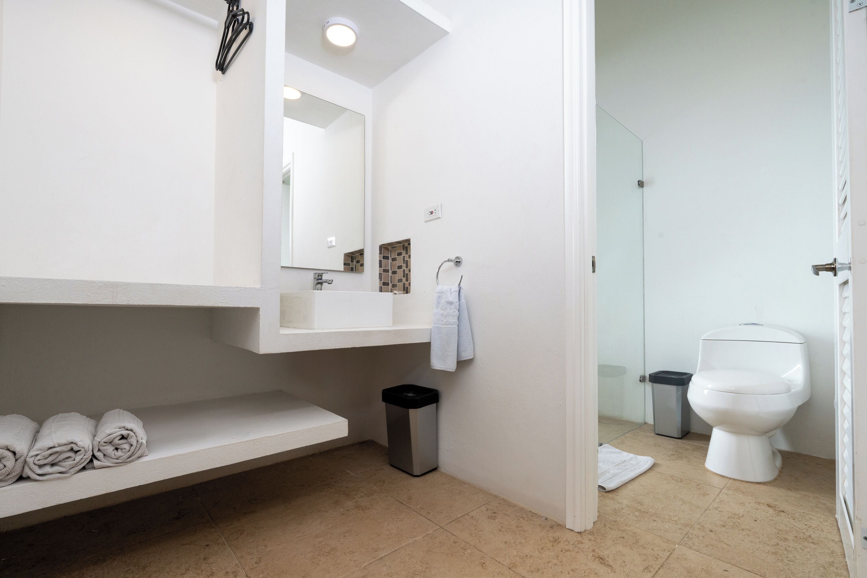 A modern bathroom with a white sink, mirror, and toilet. There is a shelf with towels and a trash bin.