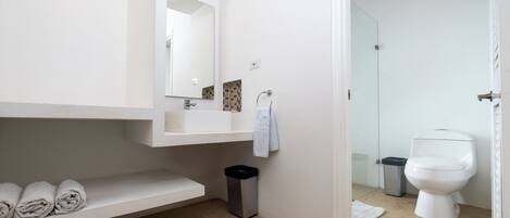 A modern bathroom with a white sink, mirror, and toilet. There is a shelf with towels and a trash bin.