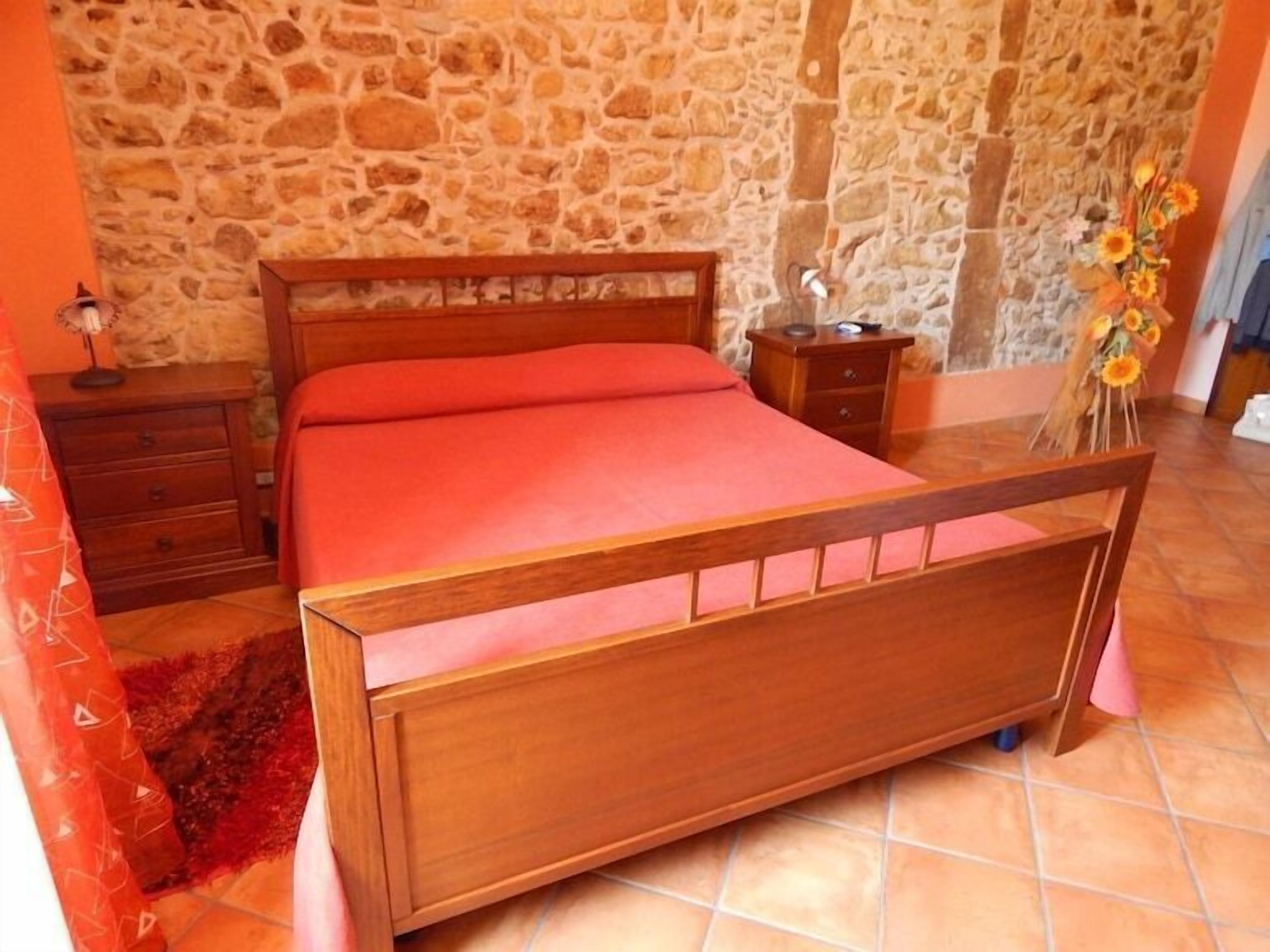 Une chambre avec un mur en pierre, un lit en bois, une table de chevet, une lampe et un vase rempli de fleurs.