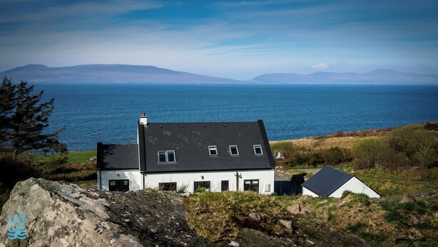 Ein Küstenhaus mit schwarzem Dach, weißen Wänden und zahlreichen Fenstern. Es liegt auf einem grasbewachsen Hügel mit Blick auf das Meer.
