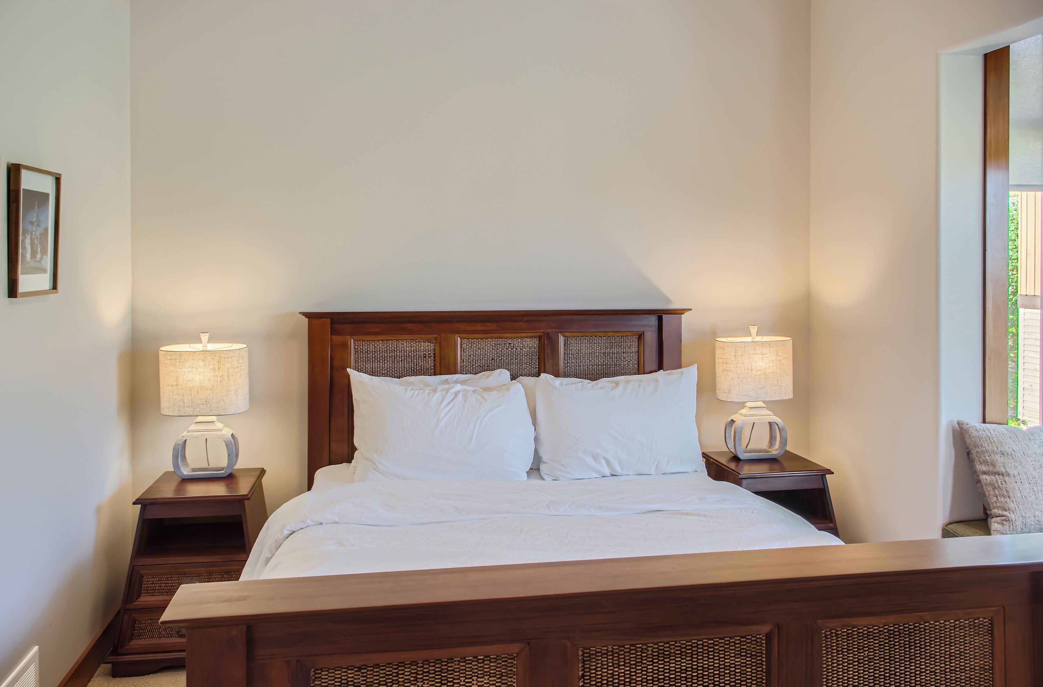 A neatly made bed with white linens, flanked by two bedside tables with lamps, in a room with a wooden headboard and a framed picture on the wall.