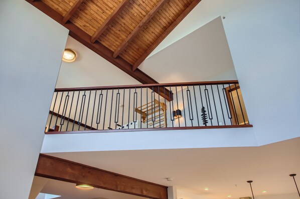 A multi-level interior with exposed wooden beams, a balcony with metal railings, and recessed lighting. - 365 Moonshine Crescent by Okanagan Premier (Beaverdell)