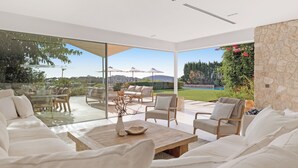 Una sala de estar moderna con pared de vidrio, muebles blancos y vista a una piscina y jardín.