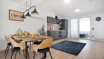 Een moderne keuken met grijze kastjes, een magnetron en een koffieapparaat. Er staat een eettafel met stoelen en een blauw vloerkleed.