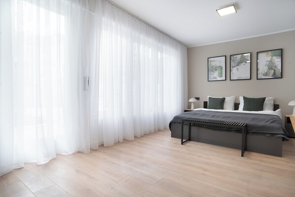 A modern bedroom with a large window covered by sheer curtains, a bed with a dark headboard, and three framed pictures on the wall. - Central Bonn Apartments I home2share (Bonn)