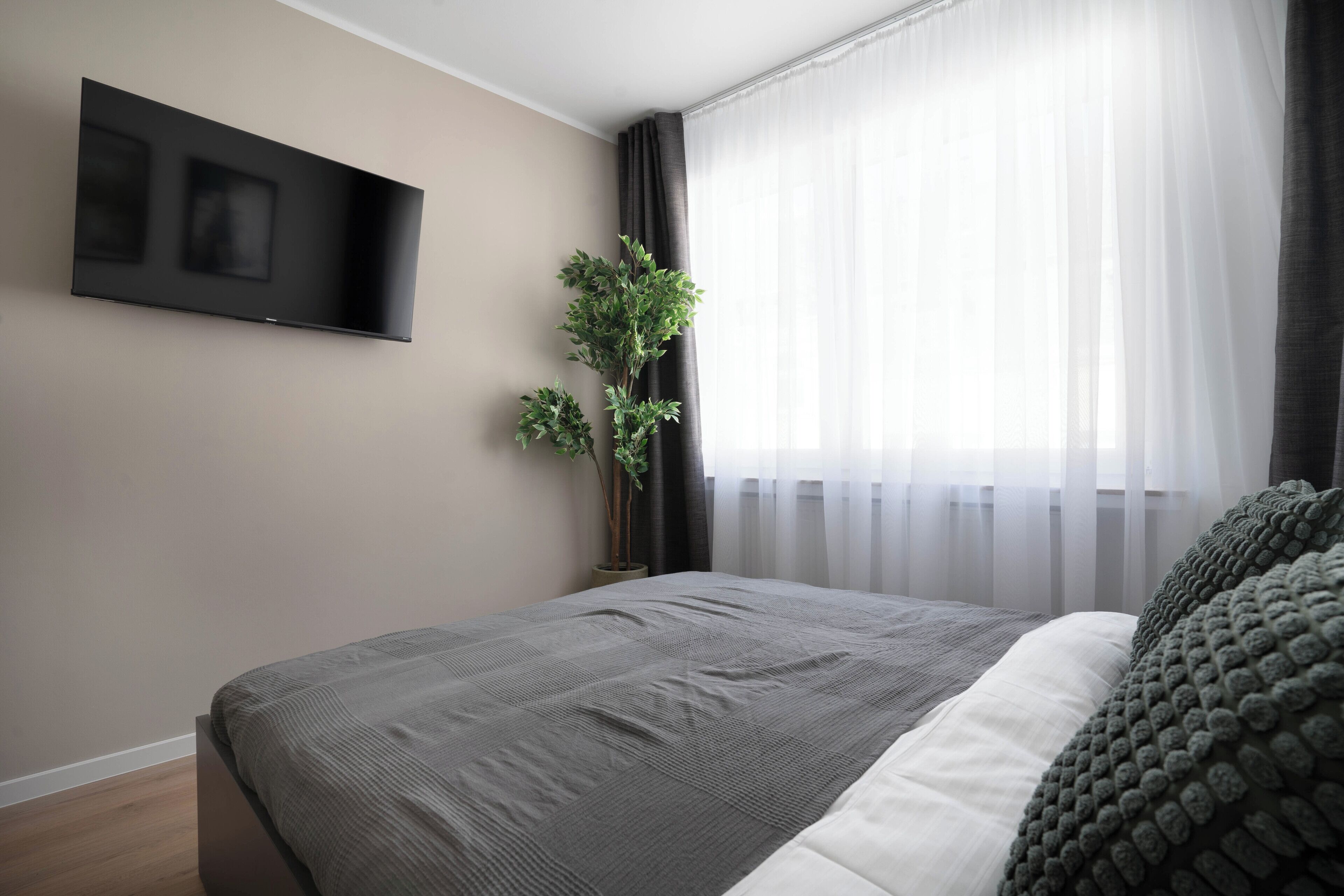 Ein modernes Schlafzimmer mit einem großen Bett, einem flachbildfernseher an der Wand, einer Topfpflanze und durchsichtigen Vorhängen am Fenster.
