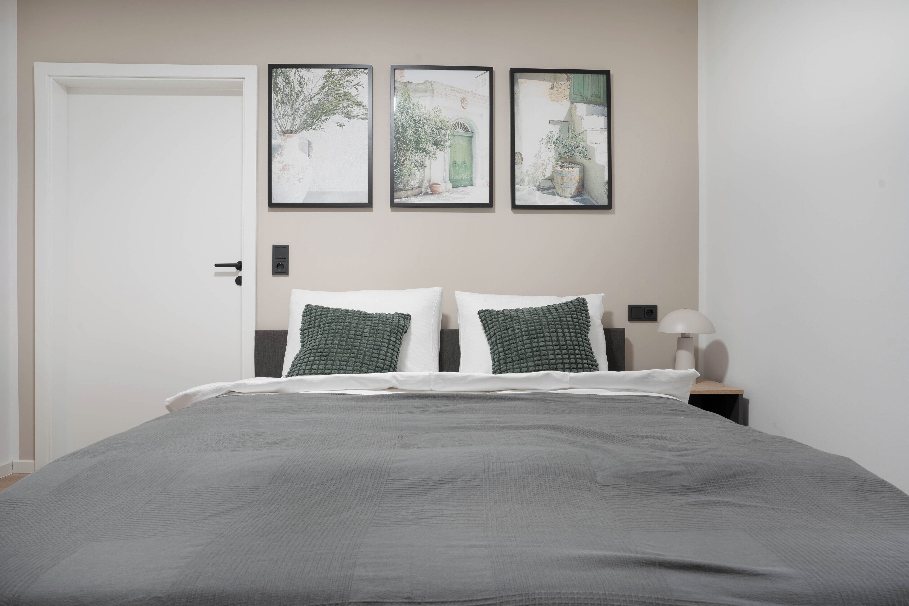 Una cama bien hecha con cubrecama gris y almohadas verdes, una mesita de noche con lámpara, y tres cuadros enmarcados en la pared.