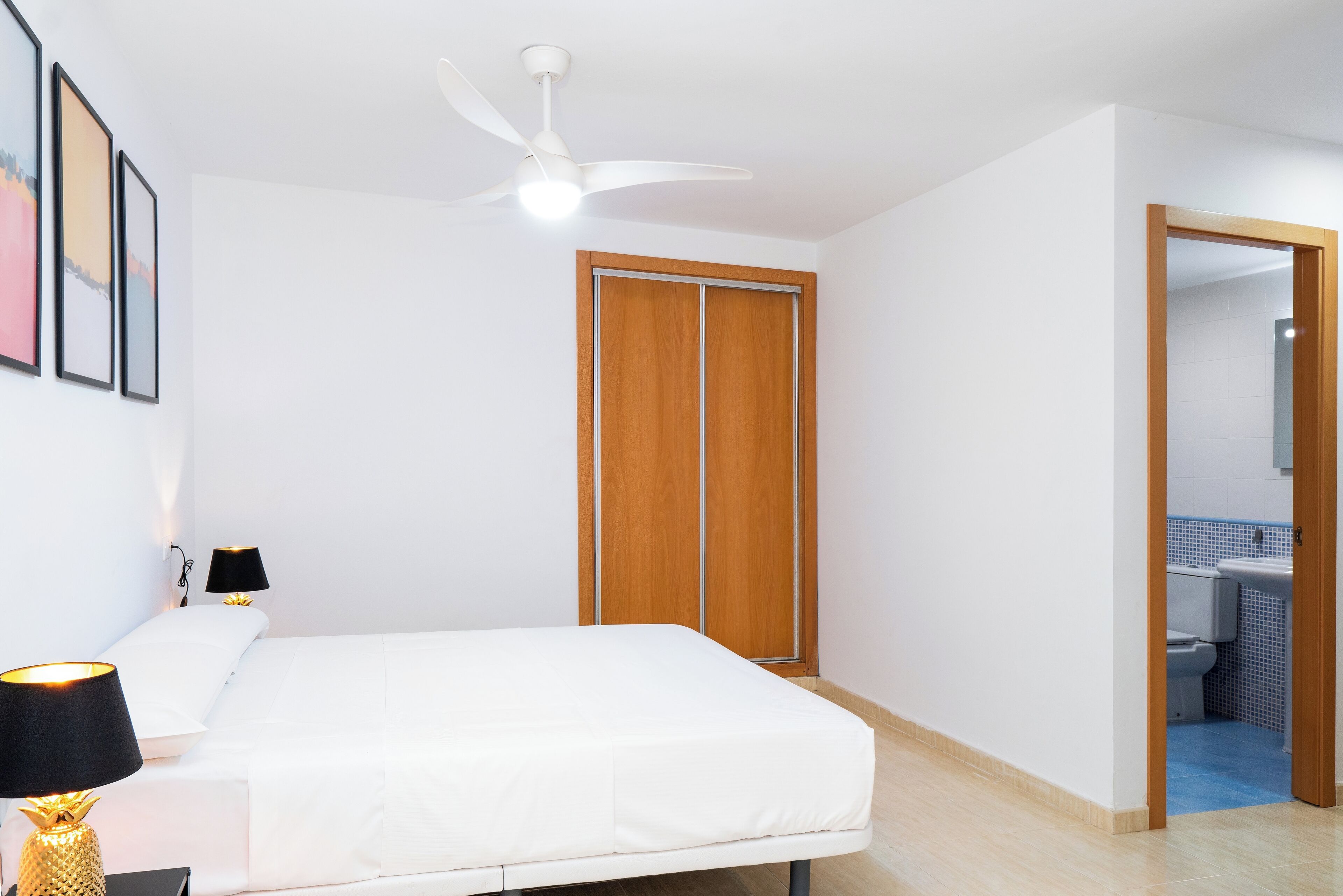 A bedroom with a bed, bedside lamps, a ceiling fan, a wooden wardrobe, and a bathroom visible through an open door.