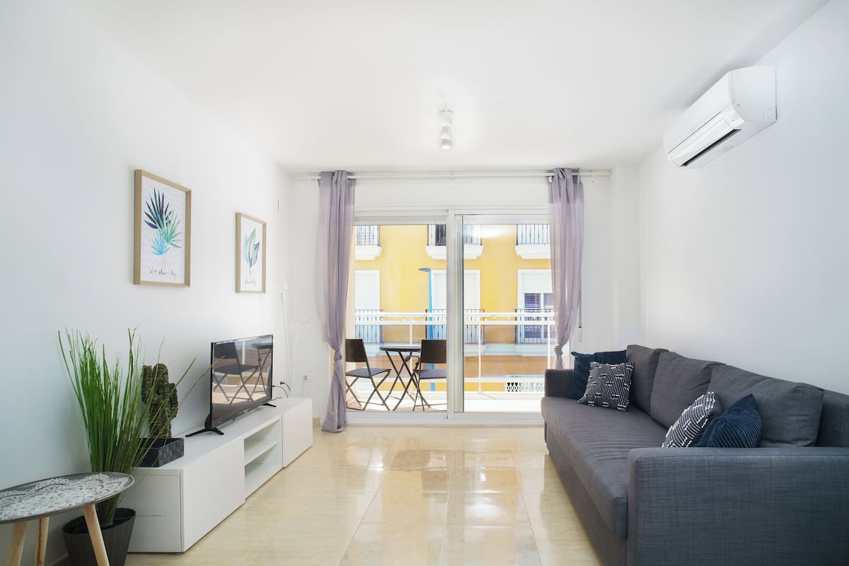 A modern living room with a grey sofa, a TV on a white stand, a small table with a plant, and a balcony with yellow buildings in the background.